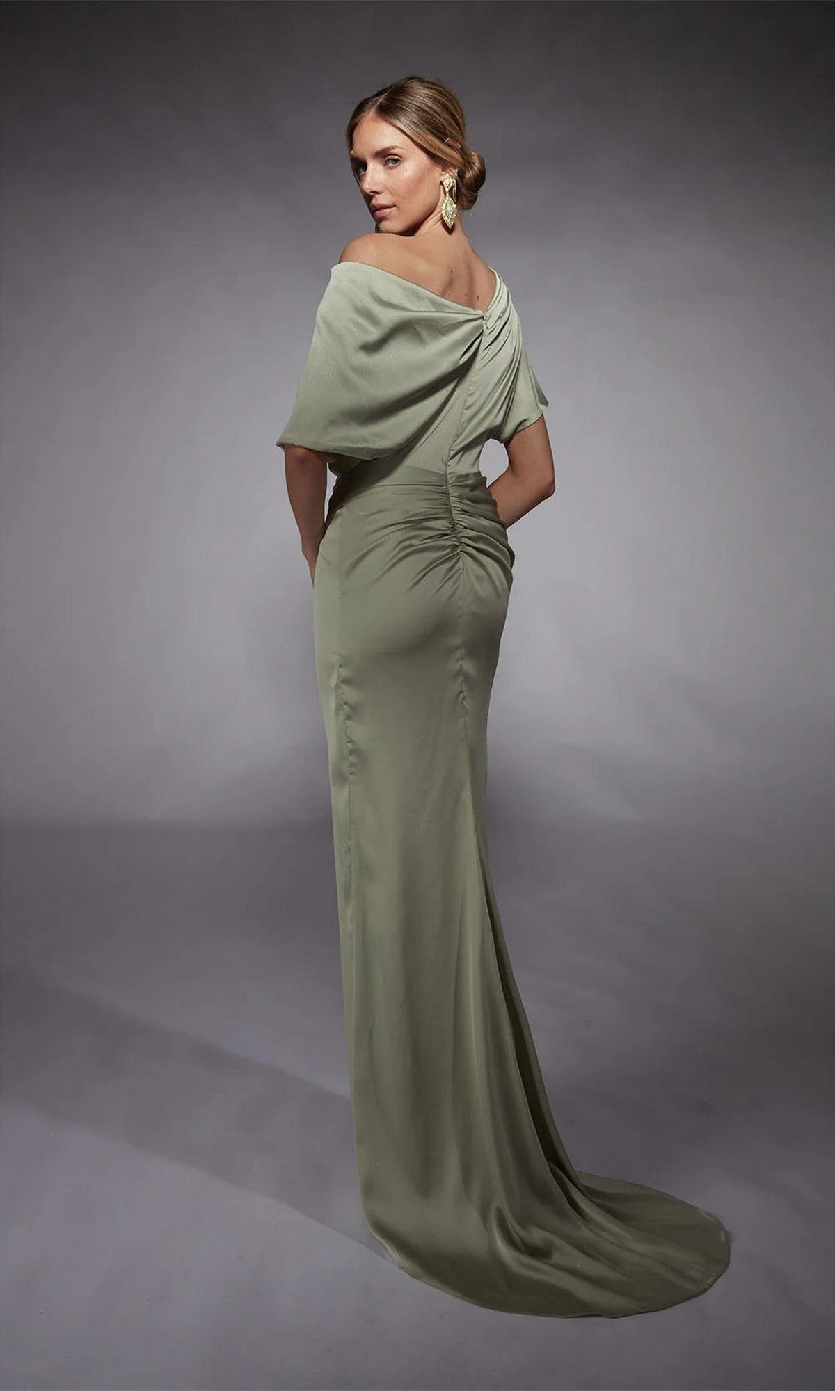 A woman poses in a studio wearing the Alyce 27748: a light sage green, off-the-shoulder formal satin dress with a ruched bodice and gathered detail at the back.